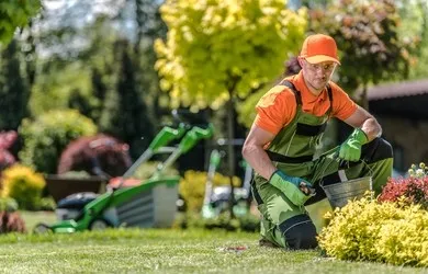 Gärtner mit Kappe pflegt ein Beet neben einem Rasenmäher